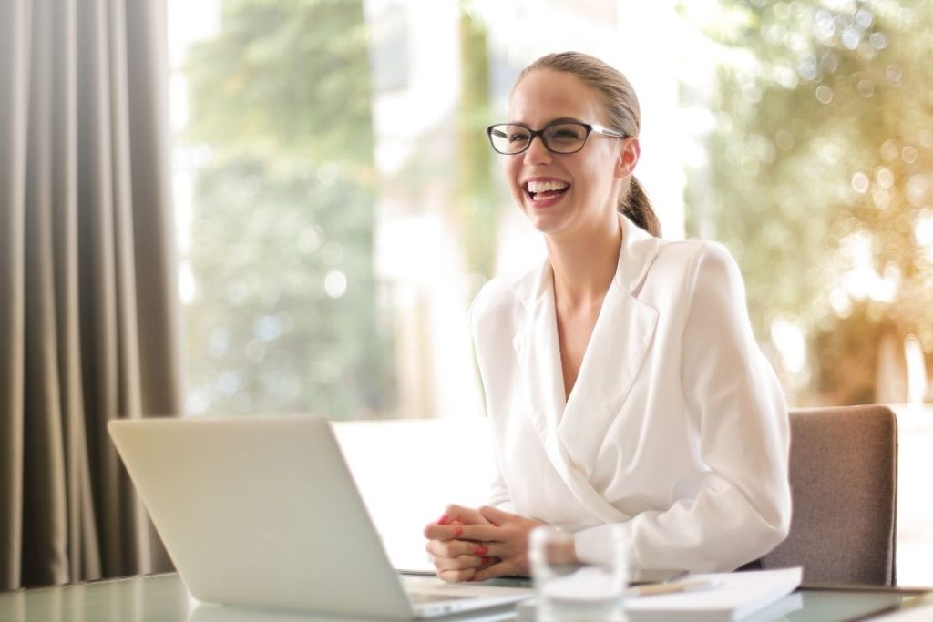 Uma mulher sorrindo, sentada em uma mesa de escritório com uma janela ao fundo com uma cortina.