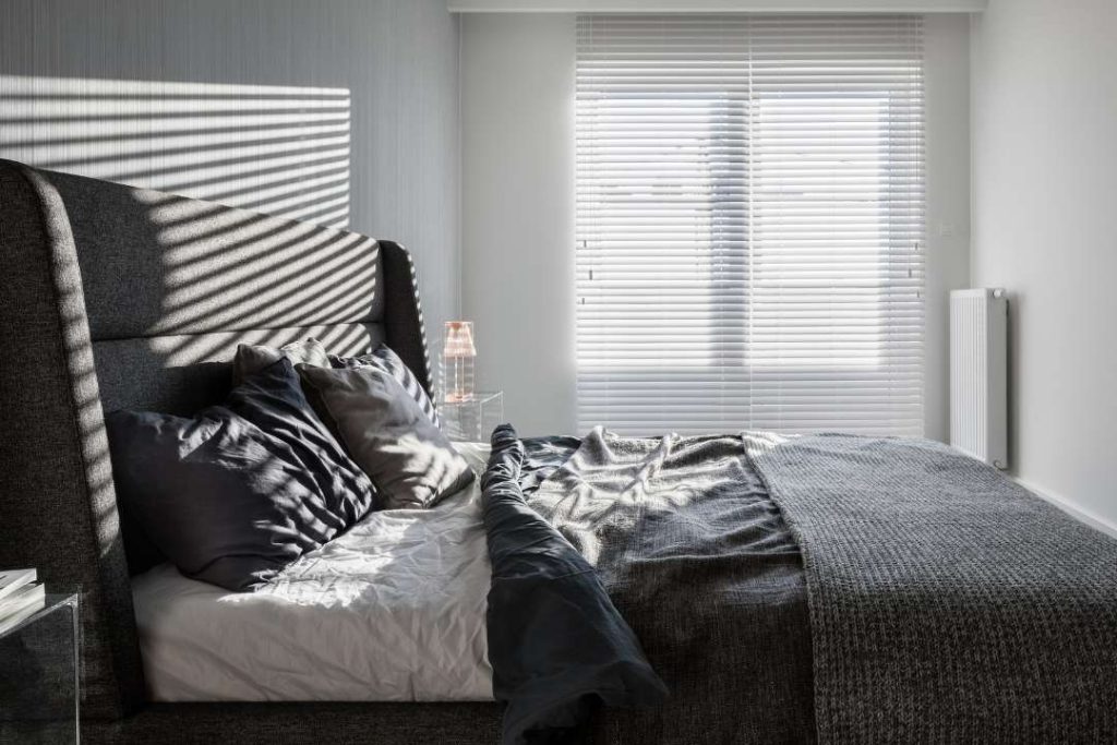 Quarto com decoração minimalista em tons de branco e cinza. As persianas brancas, parcialmente abertas, permitem a entrada suave da luz natural, criando um reflexo aconchegante no ambiente.