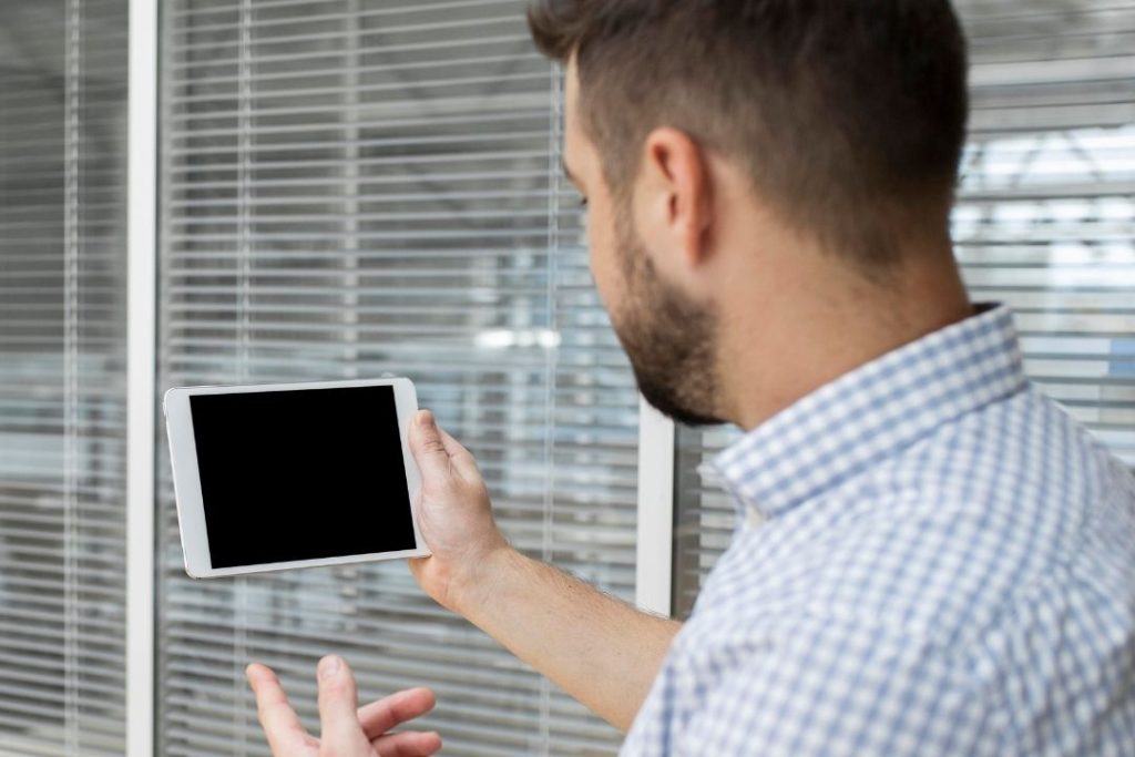 Um homem visto de costas segura um tablet com a tela preta em uma sala iluminada, na frente de janelas com persianas fechadas. 
