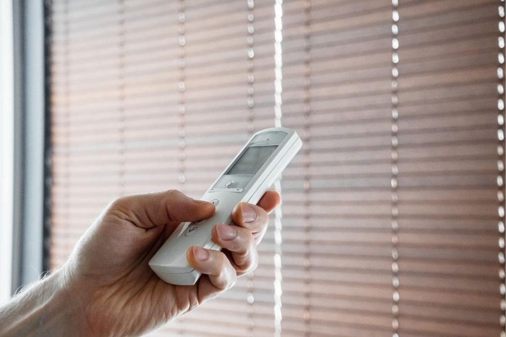 Uma mão segura um controle remoto branco apontado para persianas de madeira fechadas em um ambiente iluminado.