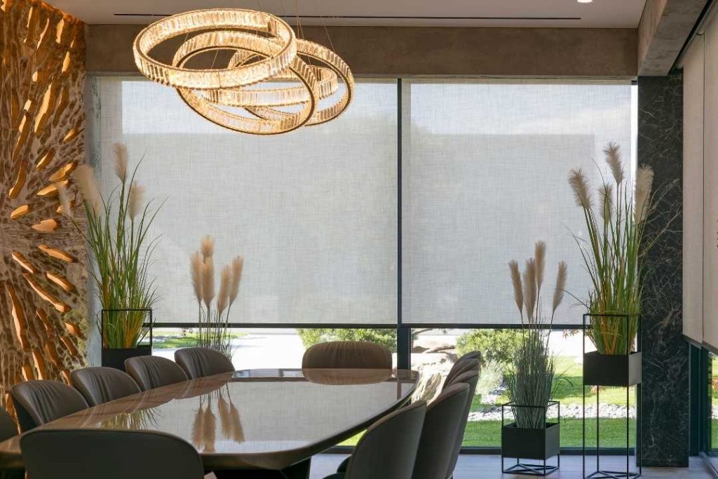 Um ambiente de sala de reunião moderno, com uma mesa de vidro, cadeiras elegantes, uma luminária suspensa dourada e grandes janelas com tela solar.