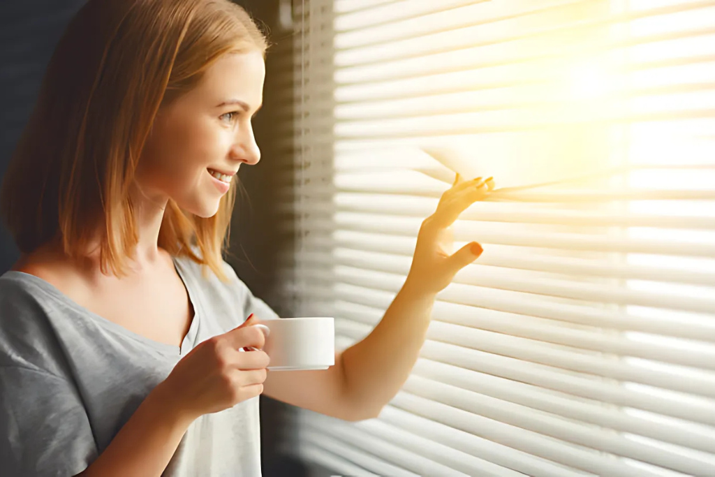 mulher observando a paisagem através da sua persiana anti calor, conseguindo evitar a luz solar e o forte calor dentro de sua casa