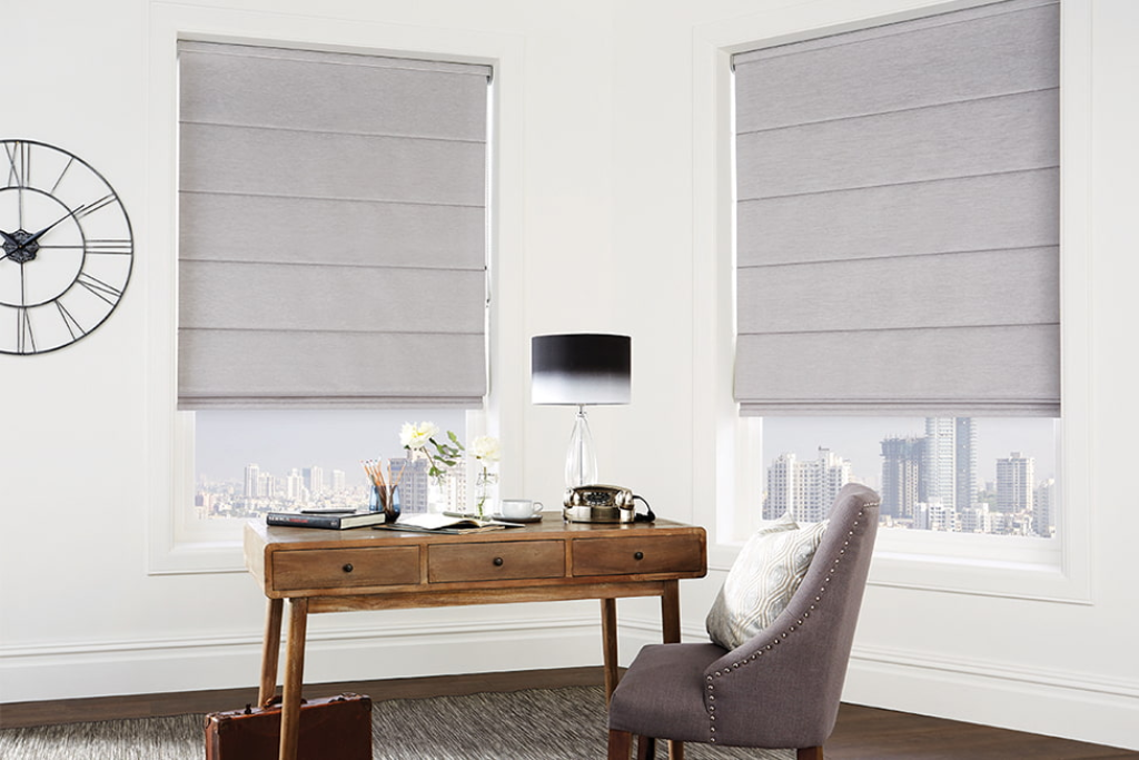 ambiente de home office com persiana romana blackout, mesa de escritório com agenda, flores, luminária, lápis e canetas, poltrona com almofada, relógio de parede e tapete